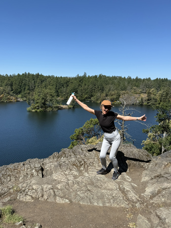 Marisa Cadot posing playfully on a rocky cliff edge overlooking a deep blue lake surrounded by a dense evergreen forest.