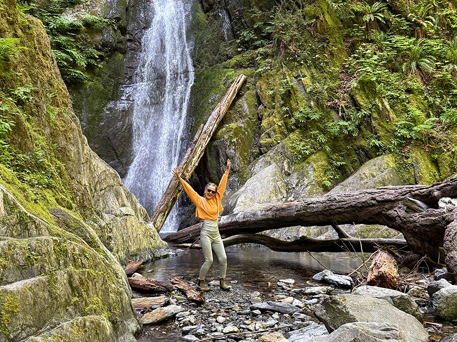 Marisa Cadot celebrating with raised arms on a rocky creek bed in front of a tall waterfall in a lush, moss-covered green forest.