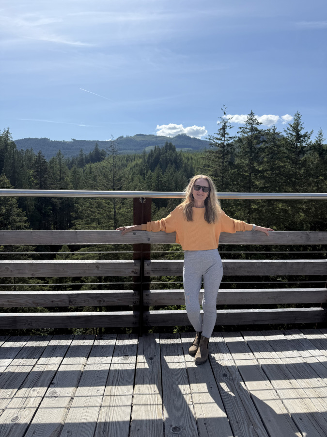 Marisa Cadot wearing a yellow sweater and sunglasses, standing on a scenic wooden boardwalk viewing platform with a vast pine forest and mountains behind her.