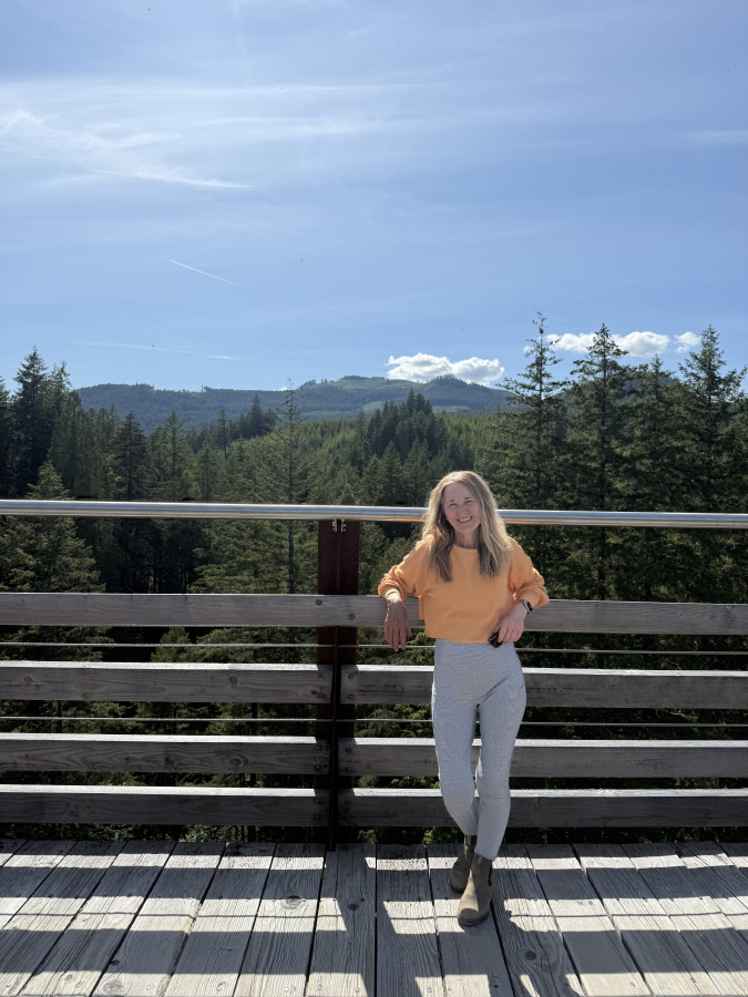 Marisa Cadot wearing a yellow sweater and sunglasses, standing on a scenic wooden boardwalk viewing platform with a vast pine forest and mountains behind her.