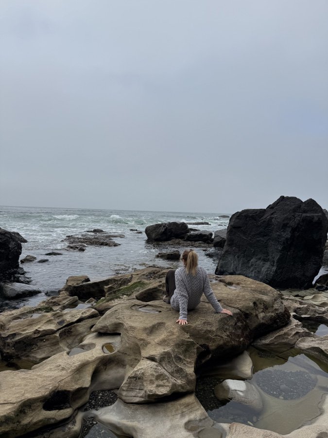 Marisa Cadot sitting on a textured rock formation overlooking a wavy ocean on an overcast day, surrounded by coastal tide pools.