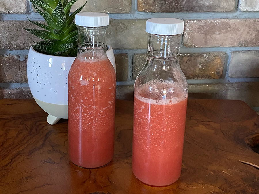 Two clear glass bottles filled with fresh, vibrant red-pink berry juice or smoothie on a wooden counter next to a potted succulent.