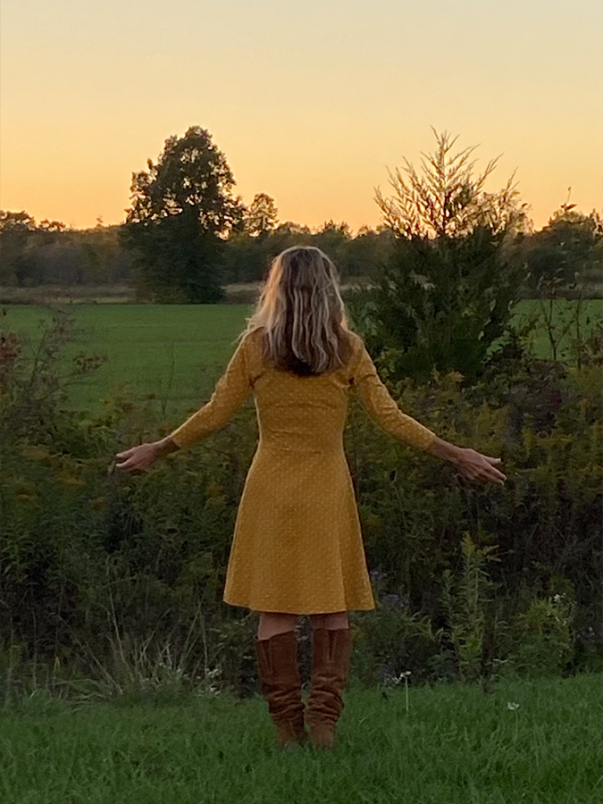 Marisa Cadot viewed from behind, standing in a grassy field with arms wide open, welcoming a glowing golden hour sunset.
