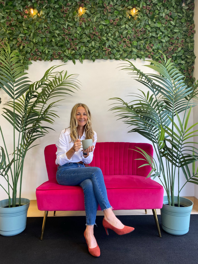 Marisa Cadot smiling while sitting on a vibrant hot pink velvet sofa holding a green coffee mug, flanked by large potted palm plants.